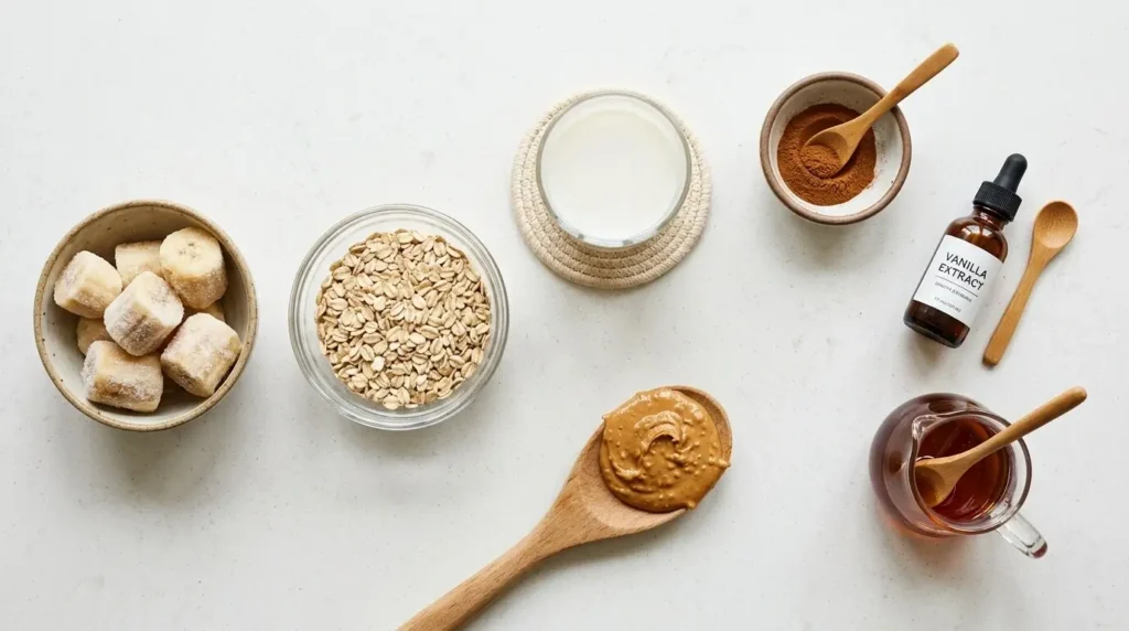 Ripe frozen banana, rolled oats, milk, cinnamon, vanilla, maple syrup and peanut butter laid out on a light countertop